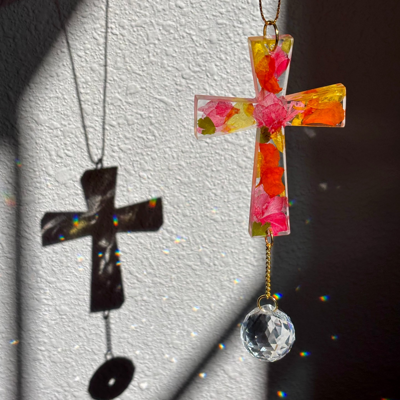 Colorful cross-shaped pendant with floral design on a chain against a white wall.
